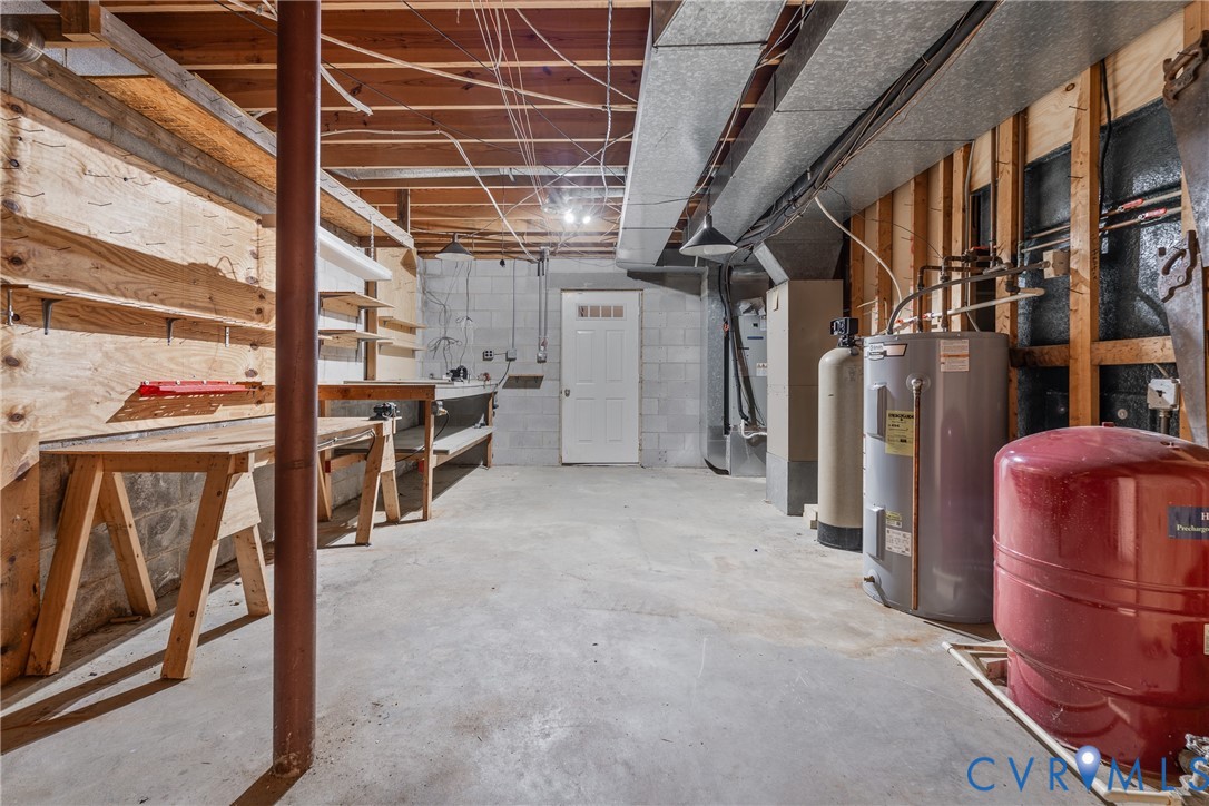 Lower-level utility area featuring exposed joist ceilings, concrete block walls, concrete flooring, built-in wooden shelving, and functional mechanical systems including a water heater and pressure tank
