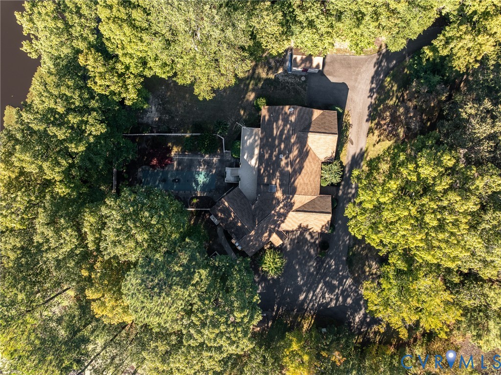Aerial perspective of a wooded lot featuring a multi-pitched roof, an asphalt driveway, and a paved patio area