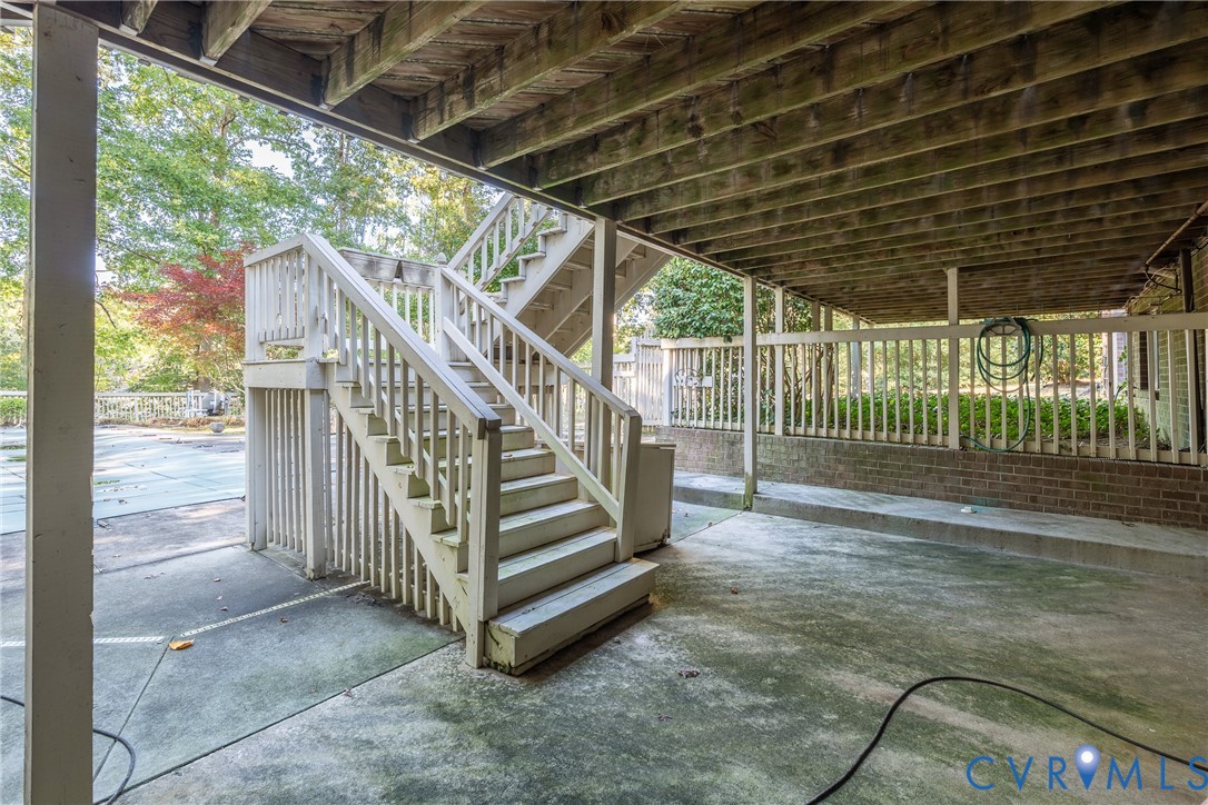 Covered patio area with a concrete floor, structural wood-beam ceiling, brick knee wall, and exterior wooden staircase with balustrades