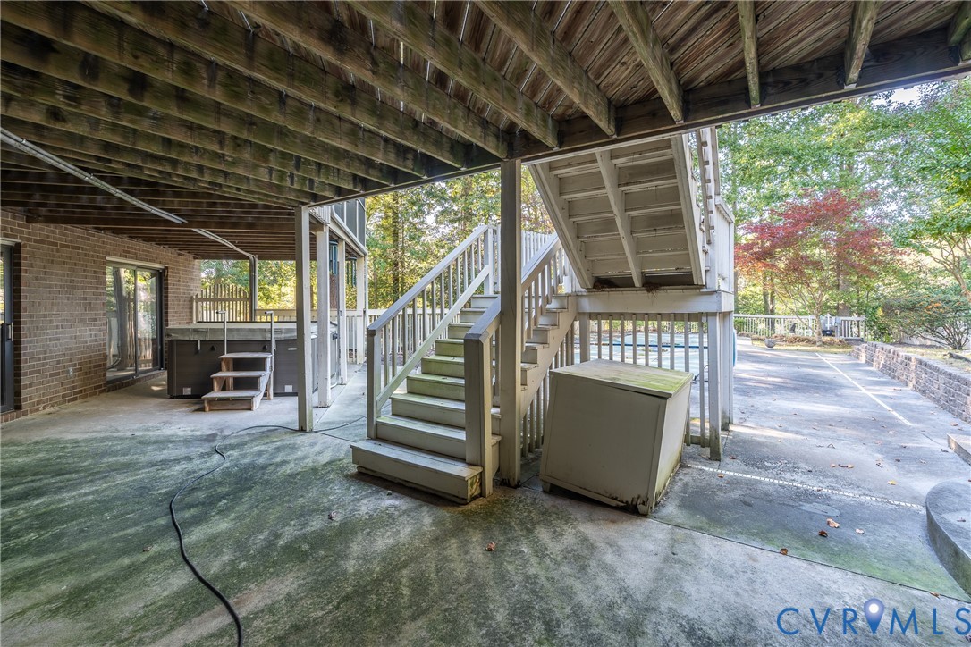 Covered concrete patio featuring brick exterior walls, wood-framed ceiling, stairs to the upper level, and a built-in hot tub