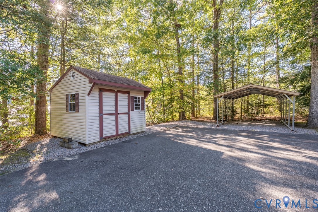 Detached storage shed with dual doors and windows, asphalt driveway, and metal-frame carport situated on a wooded lot
