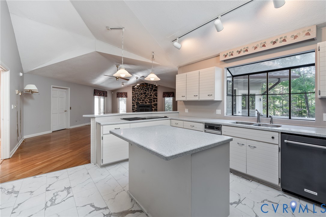 Kitchen featuring vaulted ceilings, a central island with a solid surface countertop, white horizontal-groove cabinetry, stone-clad fireplace surround, and wood-finish flooring