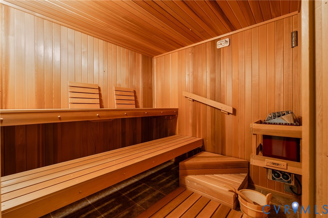 Sauna featuring wood paneling, multi-level built-in bench seating, integrated wall-mounted heater with rock basin, and wood-slat flooring