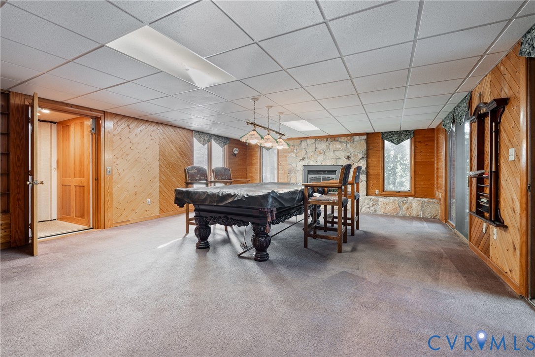 Interior space featuring diagonal wood-paneled walls, floor-to-ceiling stone fireplace surround, recessed ceiling lighting, three-light hanging fixture, and neutral carpeting