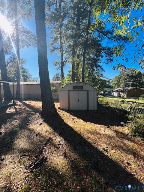Exterior storage shed with a gambrel roof, metal siding, and double-door entry situated on a wooded lot featuring mature trees and chain-link perimeter fencing