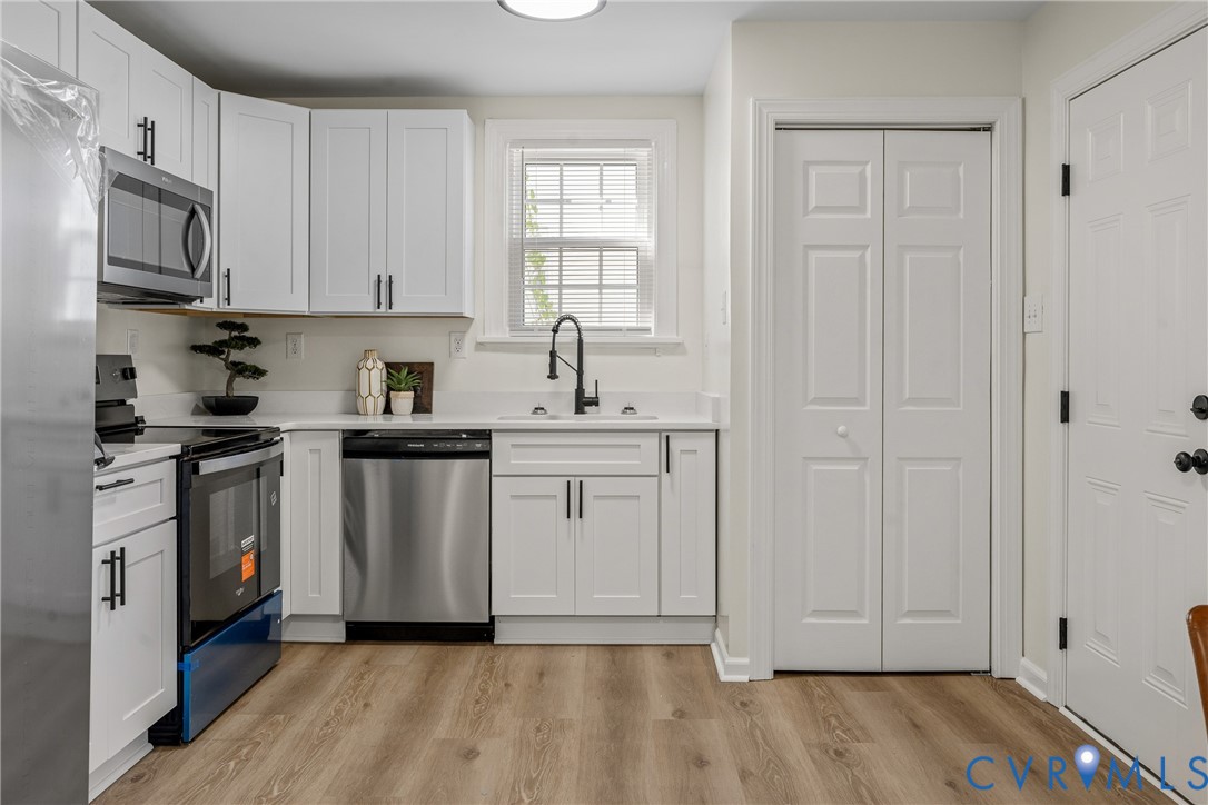 Kitchen featuring white shaker-style cabinetry, stainless steel appliances, wood-finish flooring, a matte black faucet, and a bi-fold closet Kitchen featuring white shaker-style cabinetry, stainless steel appliances, wood-finish flooring, a matte black faucet, and a bi-fold closet