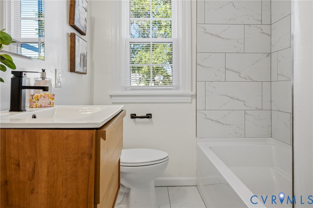 Bathroom featuring a wood-finish vanity with a white basin, matte black faucet, white tile shower surround with veining, and a window with a multi-pane sash