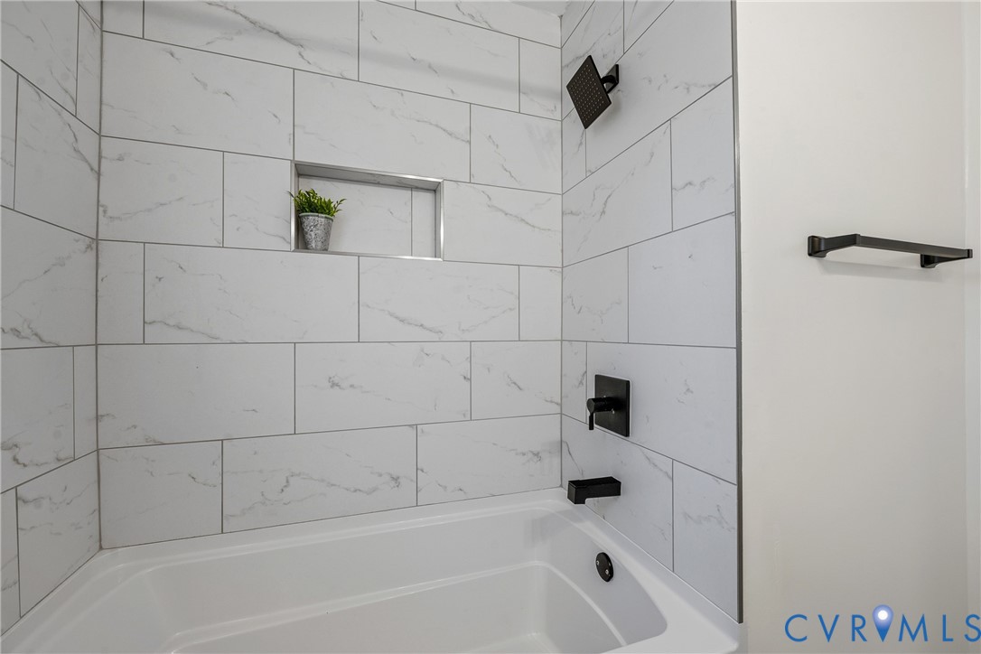 Bathtub surround featuring large-format white stone-pattern tile, a built-in recessed niche, matte black rainfall showerhead, matching matte black faucet, and a wall-mounted matte black towel bar