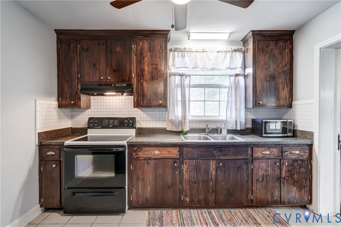 Kitchen featuring dark wood cabinetry, square tile backsplash, stone-finish countertops, stainless steel double-basin sink, and integrated overhead ceiling fan