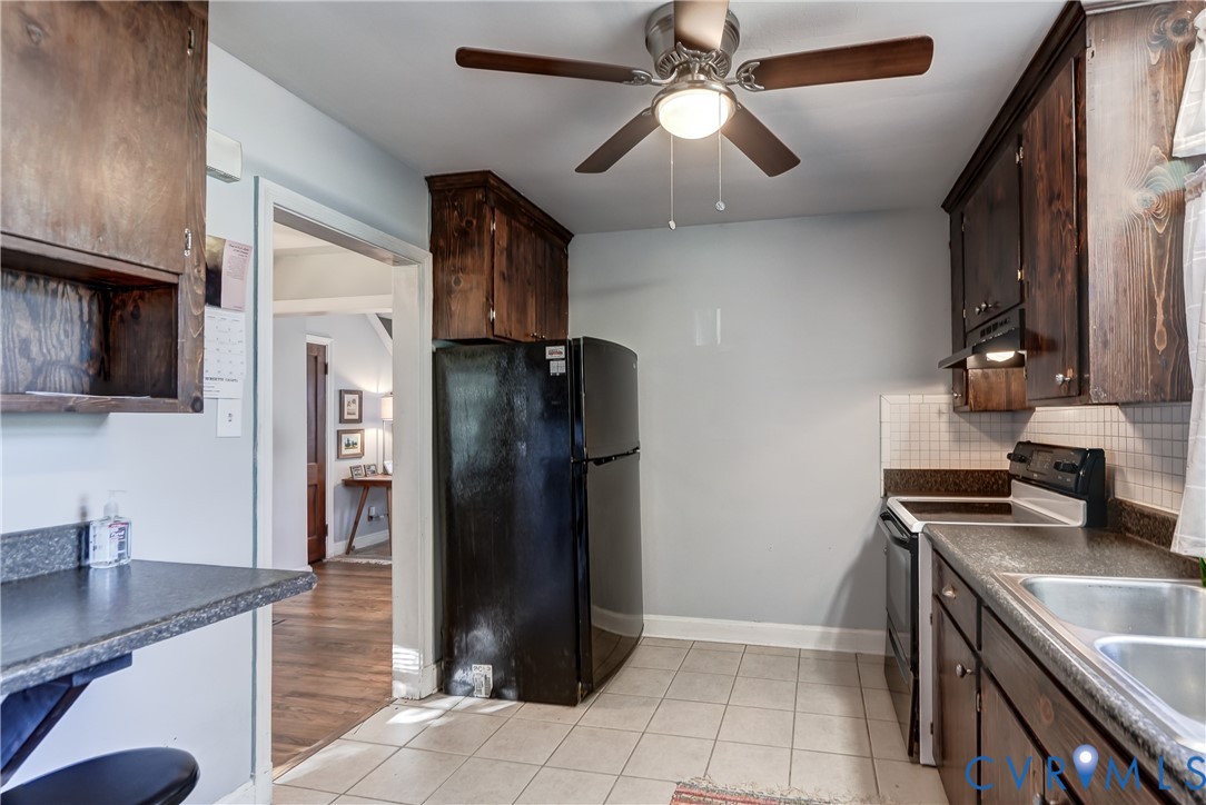 Kitchen featuring dark wood-finish cabinetry, a black refrigerator, a double-basin stainless steel sink, tiled backsplash, and a ceiling fan