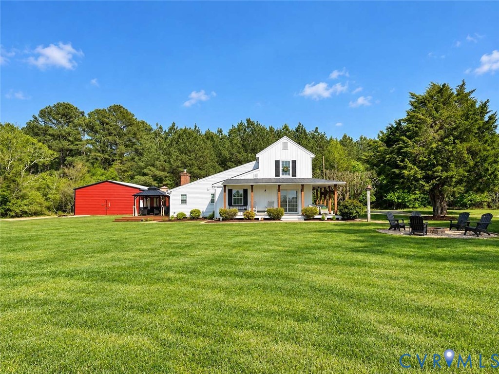 Expansive lawn featuring a white farmhouse with a covered front porch, a detached red metal barn, and a gravel fire pit area