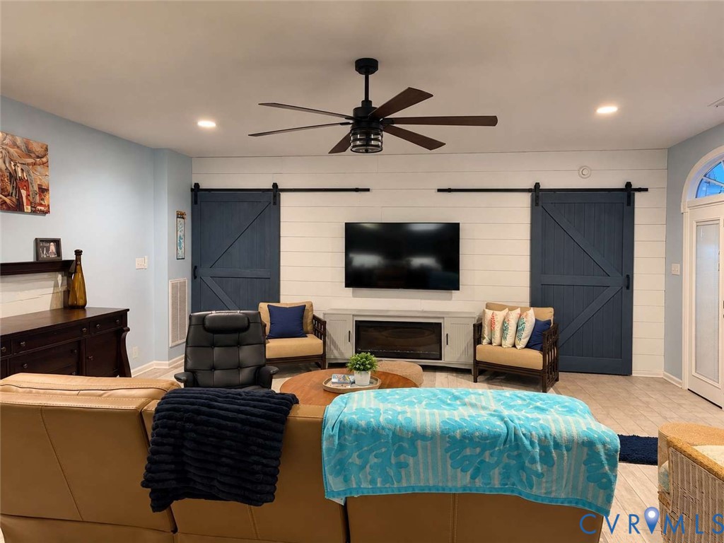 Horizontal shiplap accent wall featuring dual sliding barn doors, integrated electric fireplace, matte black ceiling fan, and light-tone tile flooring