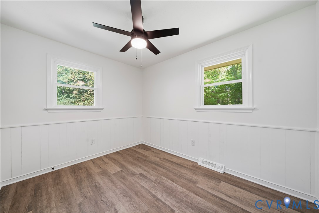 Room featuring white wainscoting, wood-finish flooring, dual windows, and a dark-finish ceiling fan with an integrated light fixture