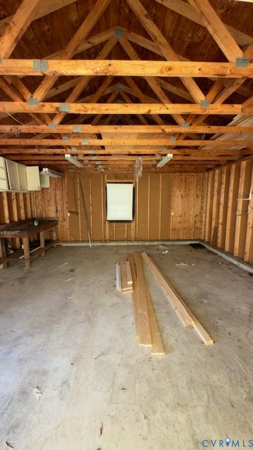 Outbuilding interior featuring exposed wood truss ceiling, concrete flooring, built-in wooden workbench, and fluorescent lighting