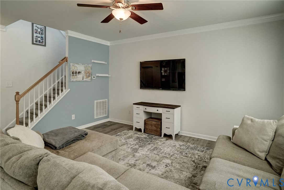 Wood-finish flooring, staircase with a wood handrail and white balusters, ceiling fan with integrated lighting, and crown molding