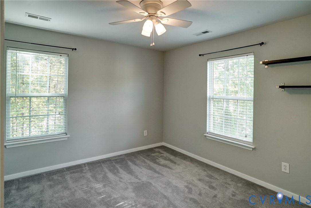 Neutral-toned interior room featuring wall-to-wall carpeting, dual windows with curtain rods, a ceiling fan with light fixture, and mounted wall shelving