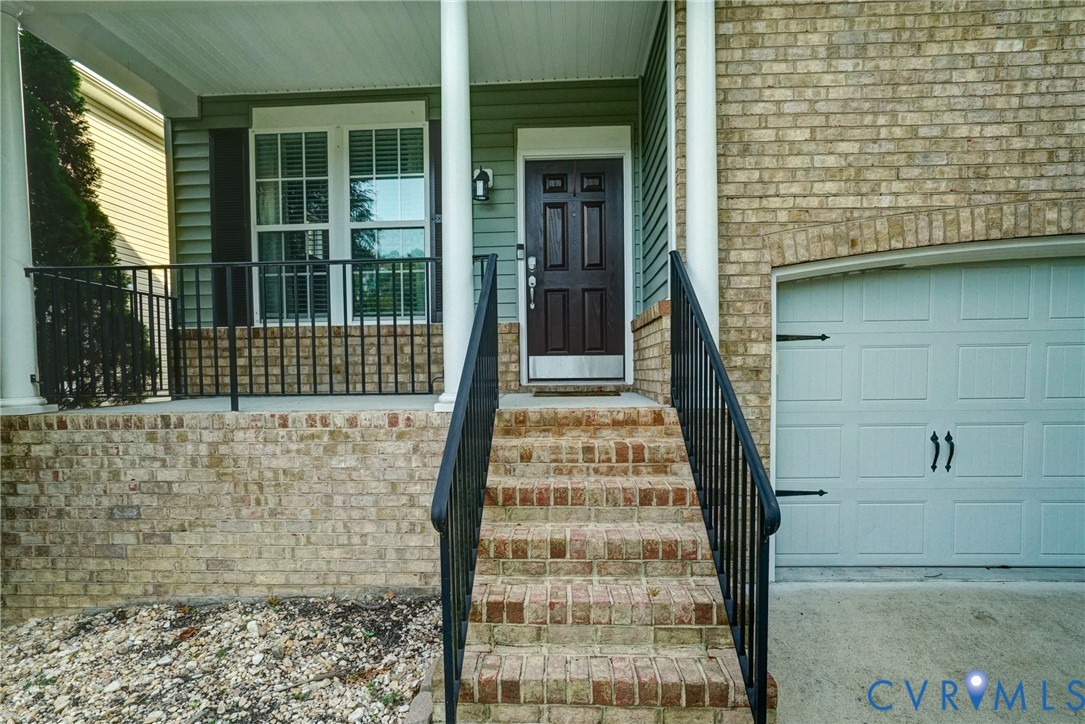 Covered front porch featuring a brick facade, decorative iron railings, and a multi-paneled entry door with a metallic kickplate, complemented by a single-car garage with a decorative exterior door Covered front porch featuring a brick facade, decorative iron railings, and a multi-paneled entry door with a metallic kickplate, complemented by a single-car garage with a decorative exterior door