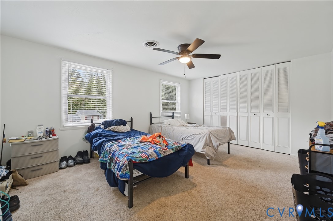 Neutral-toned room featuring wall-to-wall carpeting, multiple windows with blinds, a ceiling fan with integrated lighting, and built-in closet storage with louvered doors