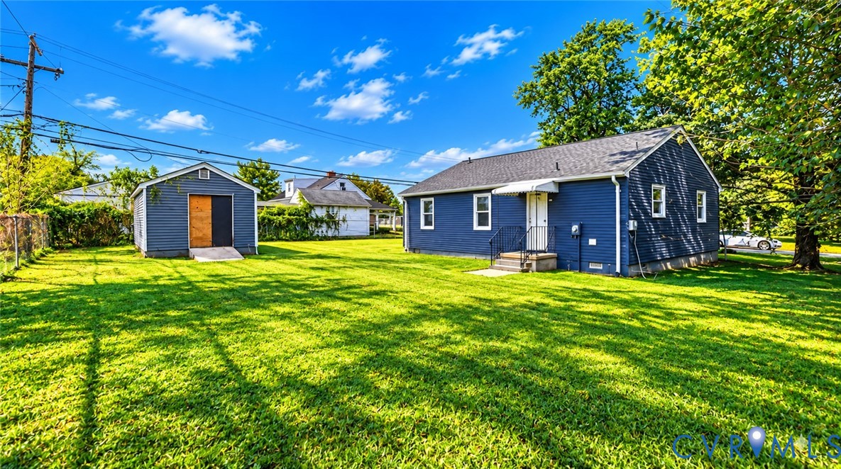 Blue-sided exterior featuring a detached storage structure, expansive lawn, metal awning, and mature trees