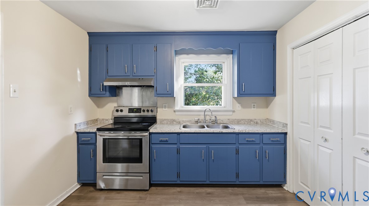 Kitchen featuring blue cabinetry, light-toned stone-finish countertops, stainless steel range with overhead vent, double-basin sink with chrome faucet, and wood-finish flooring