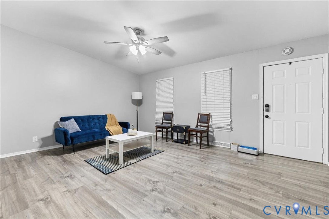 Neutral-toned interior space featuring wood-finish flooring, a multi-blade ceiling fan with integrated lighting, two windows with horizontal blinds, and a white six-panel entry door Neutral-toned interior space featuring wood-finish flooring, a multi-blade ceiling fan with integrated lighting, two windows with horizontal blinds, and a white six-panel entry door