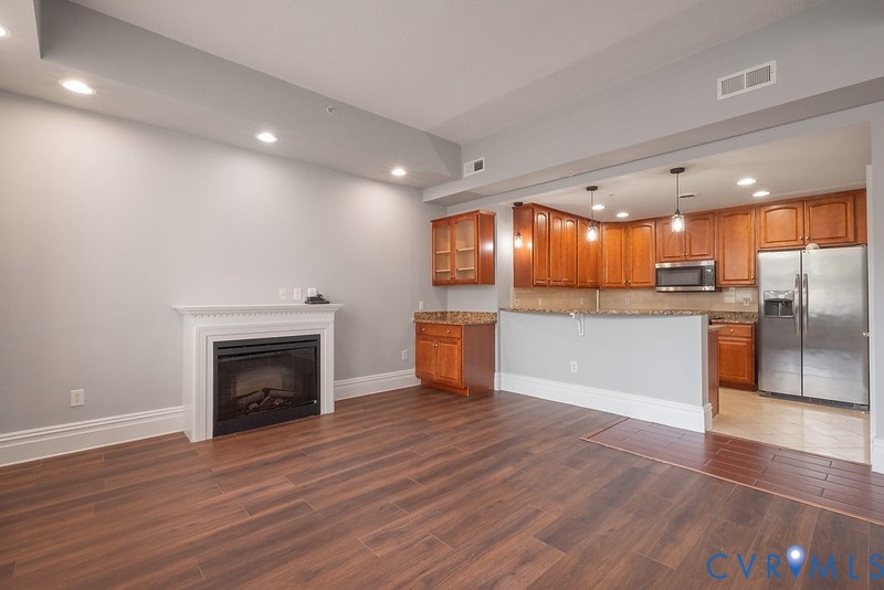 Living space featuring wood-finish flooring, a white mantel fireplace, recessed lighting, and a kitchen equipped with wooden cabinetry and a stone-surface breakfast bar