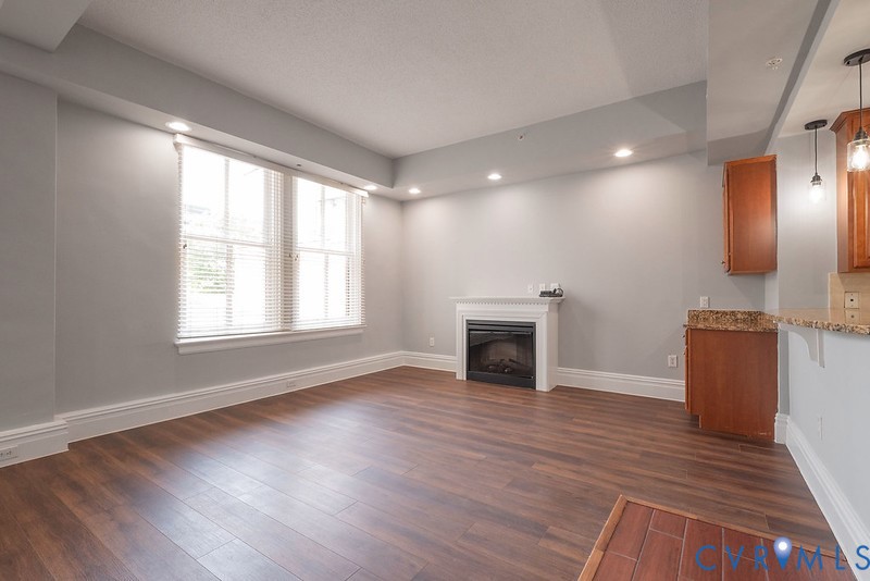Living area featuring wood-finish flooring, a recessed ceiling with integrated lighting, a fireplace with a white mantel, and wood cabinetry with stone-finish countertops
