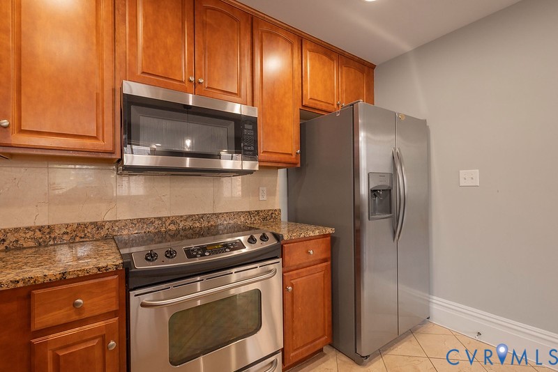 Kitchen featuring wood cabinetry, stone countertops, stone tile backsplash, stainless steel appliances, and tile flooring