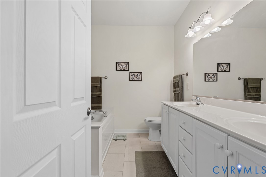 Dual-sink vanity with white cabinetry, solid-surface countertop, full-width wall mirror, four-bulb overhead vanity lighting, and ceramic tile flooring
