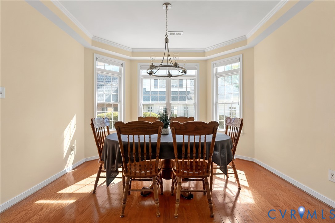 Dining space featuring bay windows, wood-finish flooring, tray ceiling, crown molding, and a tiered metal chandelier Dining space featuring bay windows, wood-finish flooring, tray ceiling, crown molding, and a tiered metal chandelier