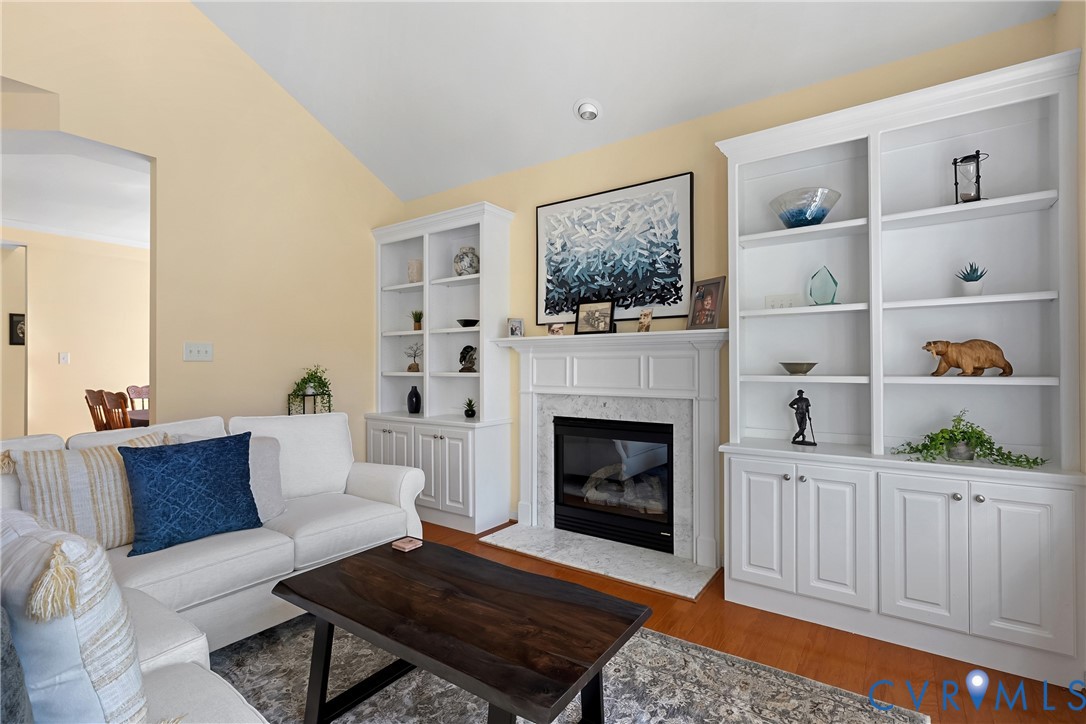 Fireplace with a stone surround and mantel, flanked by white custom built-in cabinetry and shelving, finished with wood flooring and a vaulted ceiling