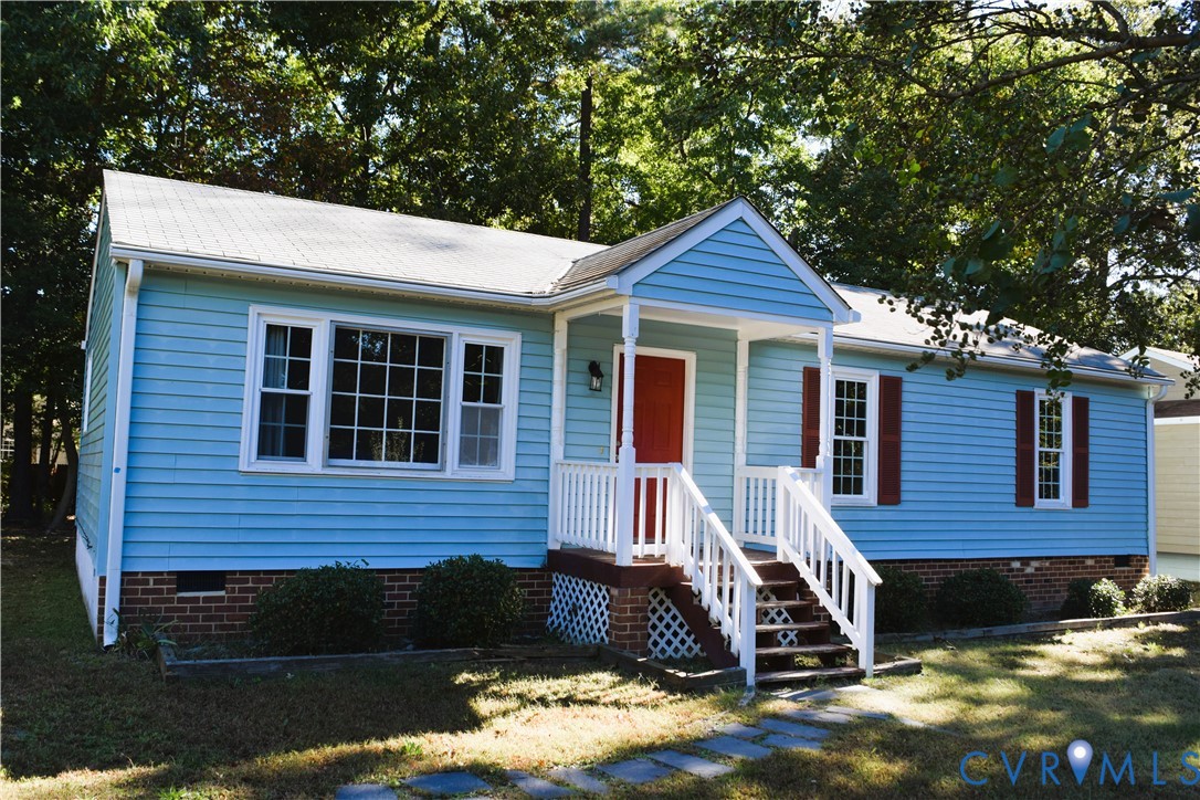 Single-story exterior featuring light blue horizontal siding, red shutters, a gable-roofed front porch, white railings, and brick foundation masonry
