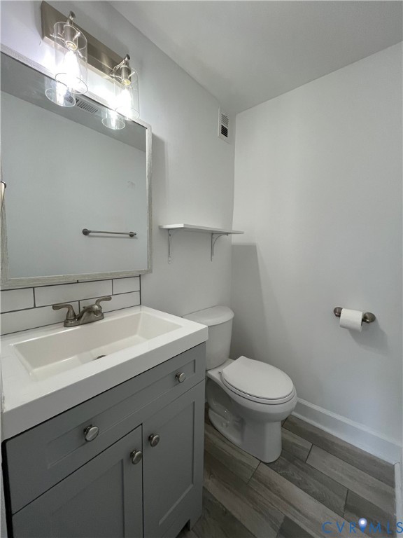 Grey vanity with integrated rectangular sink, wood-finish flooring, subway tile backsplash, wall-mounted shelving, and dual-light vanity fixture