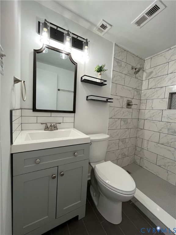 Bathroom featuring a grey vanity with a rectangular sink, stone-finish shower surround with recessed shelving, dark tile flooring, and a framed vanity mirror with three-light fixture