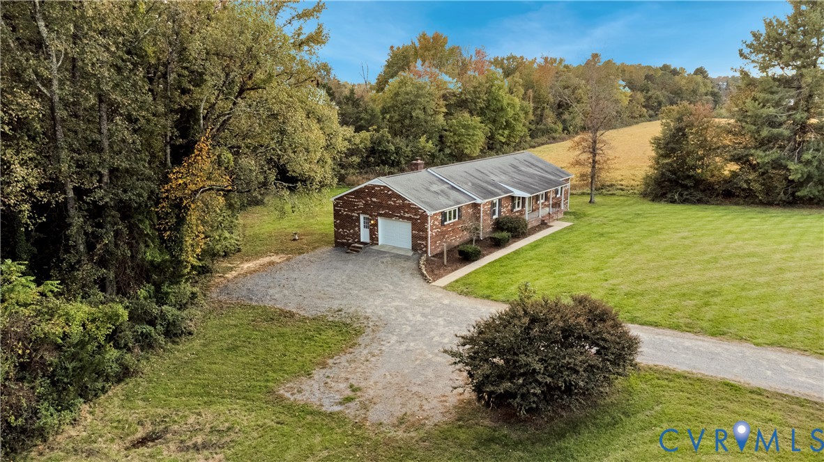 Single-story brick exterior featuring a shingled roof, an attached garage, and a gravel driveway situated on a lot with mature trees and expansive lawn space