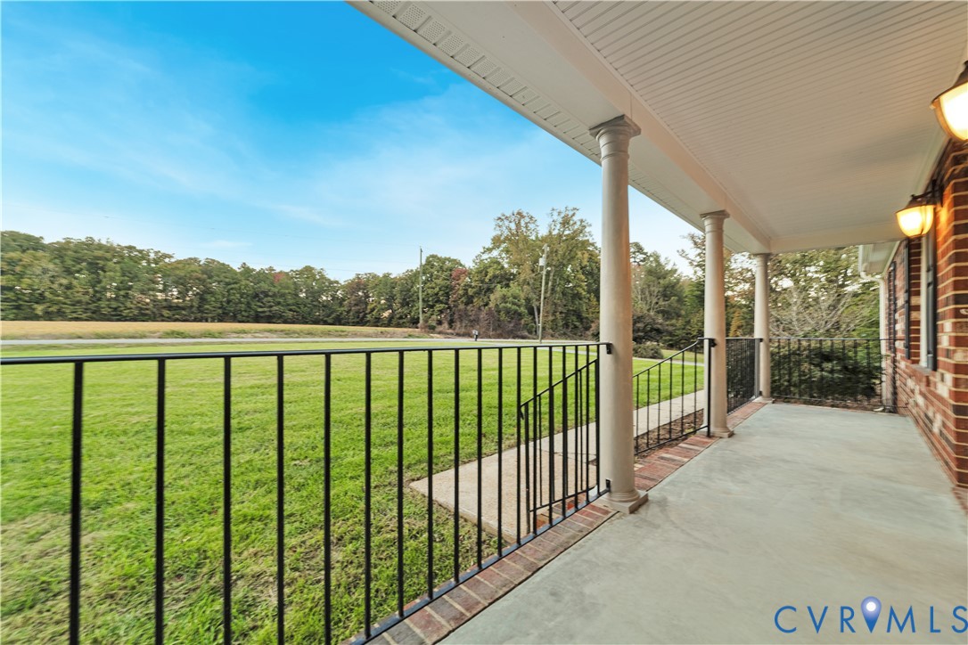 Covered front porch featuring brick-trimmed concrete flooring, classical columns, metal railing, and wall-mounted exterior lighting