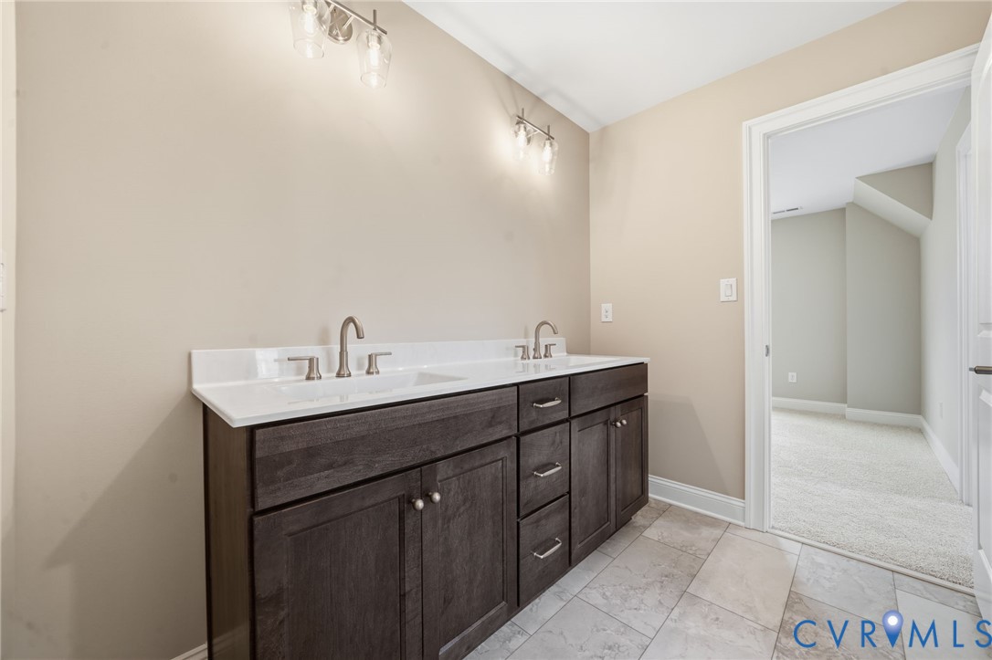 Dual-sink vanity with dark wood-finish cabinetry, solid surface countertop, metallic hardware, and wall-mounted light fixtures, complemented by tile flooring