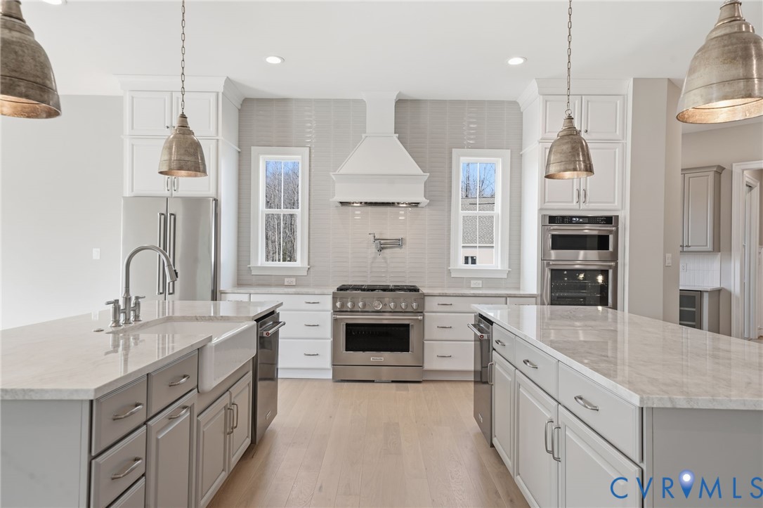 Kitchen featuring dual islands with stone-composite countertops, shaker-style cabinetry, stainless steel professional-grade appliances including a wall-mounted pot filler, and wood-finish flooring