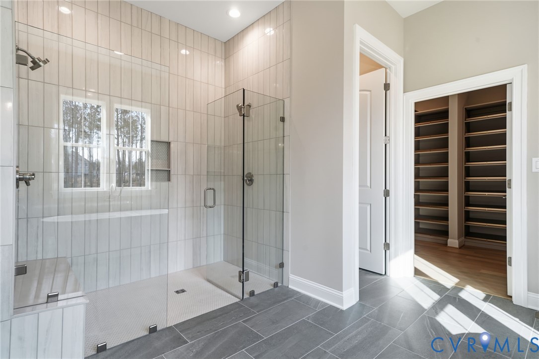 Walk-in shower featuring floor-to-ceiling vertical tile surround, glass enclosure, built-in tiled bench, and rainfall showerhead, complemented by large-format slate-finish floor tile and walk-in closet with built-in shelving