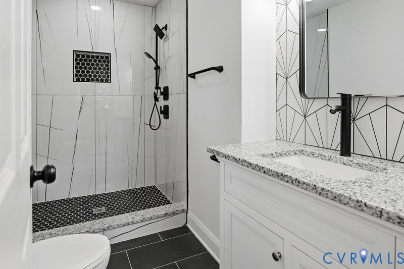 Bathroom featuring a walk-in shower with large-format tile, a stone-finish countertop with an integrated sink, matte black plumbing fixtures, and geometric tile backsplashes