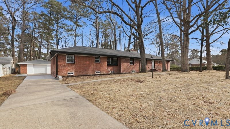 Red brick exterior featuring a hip roof, concrete driveway, detached single-car garage, and mature trees on the lot