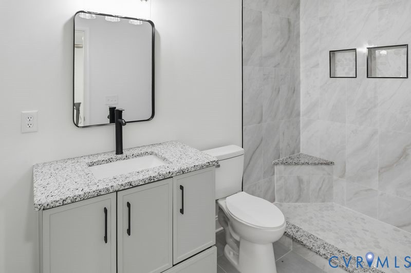 Bathroom featuring a light-toned vanity with a speckled countertop, matte black faucet, rectangular mirror, and walk-in shower with stone-tile surround, built-in bench, and recessed shelving