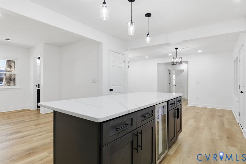 Kitchen island featuring a quartz countertop, dark-toned cabinetry, built-in beverage refrigerator, and trio of pendant light fixtures, complemented by wood-finish flooring