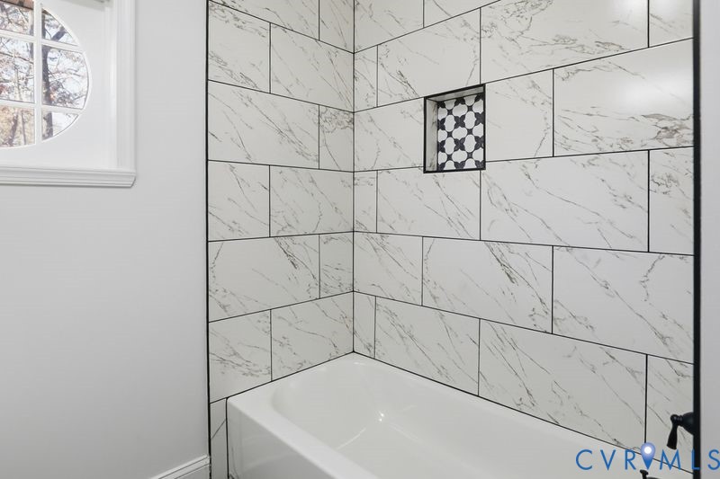 Bathtub surround featuring large-format white veined wall tile, a recessed niche with patterned accent tile, matte black fixtures, and an integrated circular window