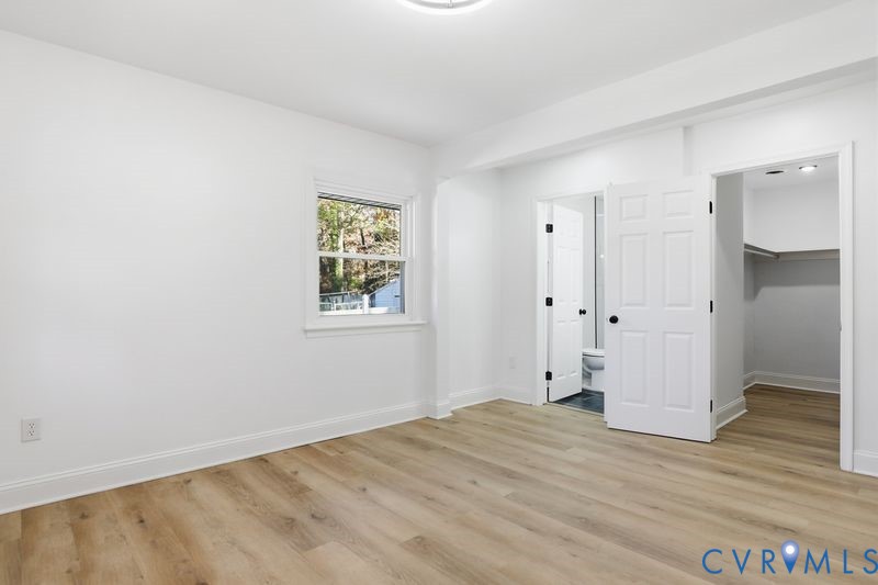 Room featuring wood-finish flooring, white walls, a multi-pane window, and two paneled interior doors opening into a bathroom and a walk-in closet