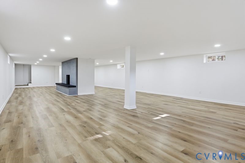 Open-concept interior featuring wood-finish flooring, recessed lighting, and a prominent center fireplace with a dark brick surround and built-in hearth