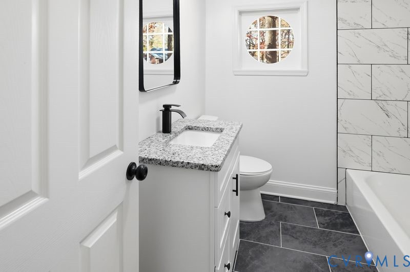 Bathroom featuring a white vanity with a stone countertop, a circular accent window, a bathtub with tiled surround, matte black hardware, and dark tile flooring