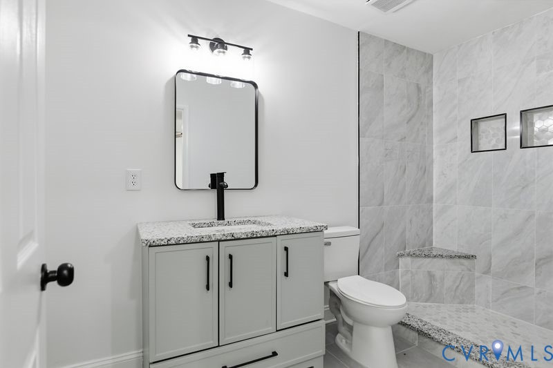 Bathroom featuring a light gray vanity with a speckled countertop, black hardware, a rectangular vanity mirror, and a tiled shower stall with built-in storage niches and a stone-topped bench