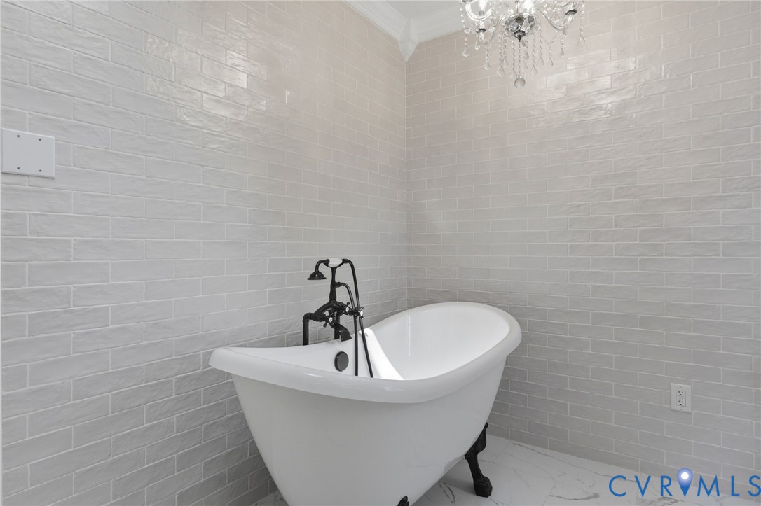 Freestanding clawfoot tub featuring a black faucet assembly, floor-to-ceiling textured tile wall, crown molding, and crystal chandelier
