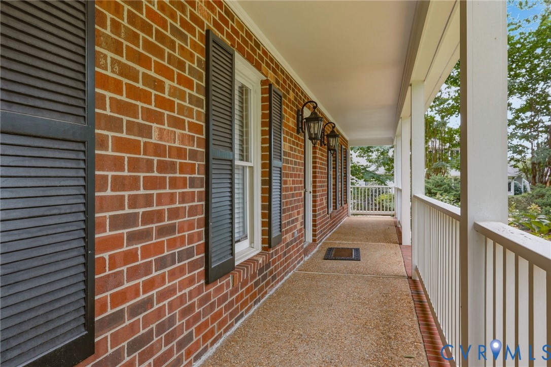 Covered front porch featuring red brick exterior wall, dark louvered shutters, exposed aggregate concrete flooring, and metal carriage-style wall lanterns Covered front porch featuring red brick exterior wall, dark louvered shutters, exposed aggregate concrete flooring, and metal carriage-style wall lanterns